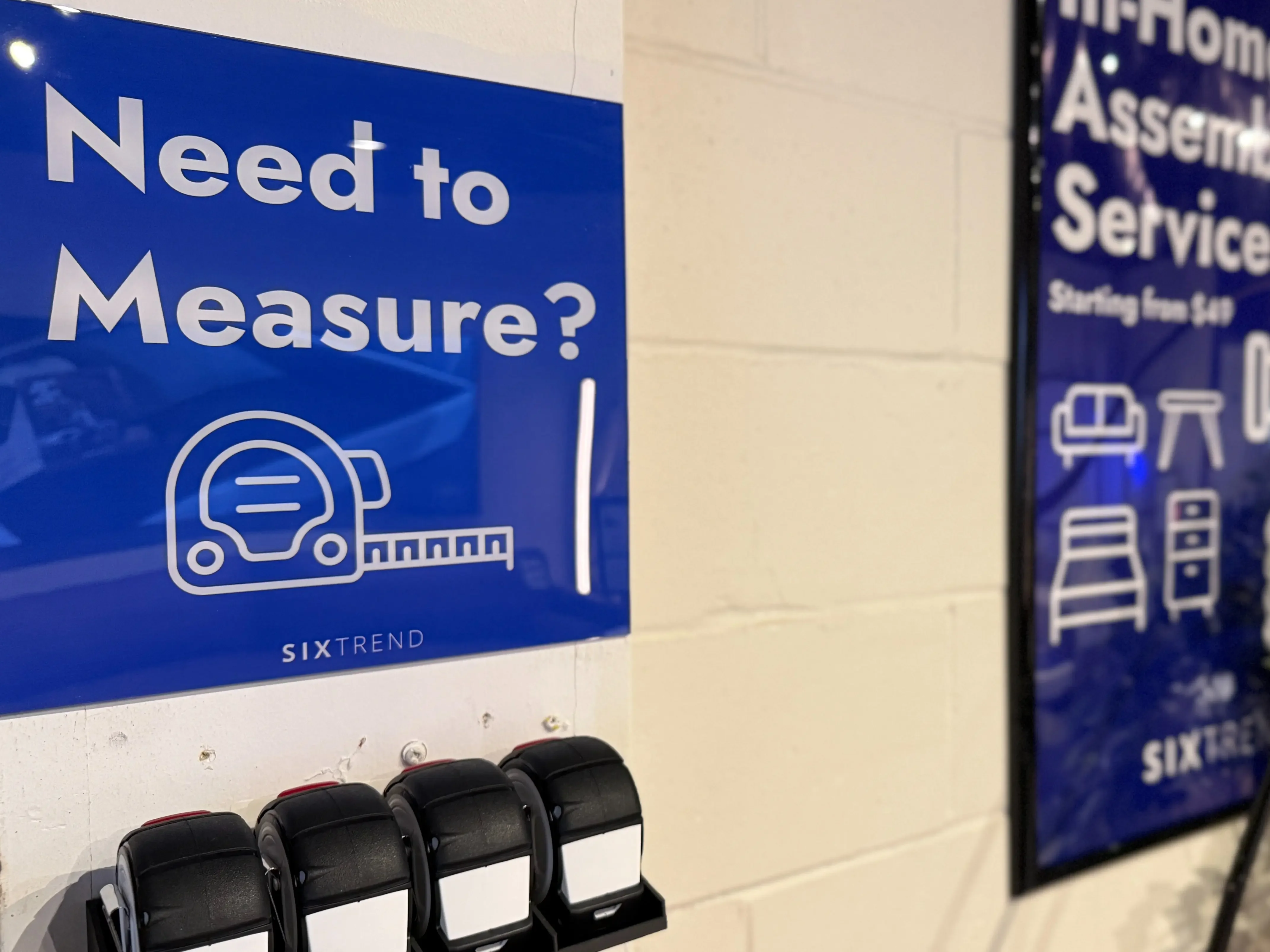 A blue 'Need to Measure?' SIXTREND sign mounted in the showroom above a row of complimentary tape measures.