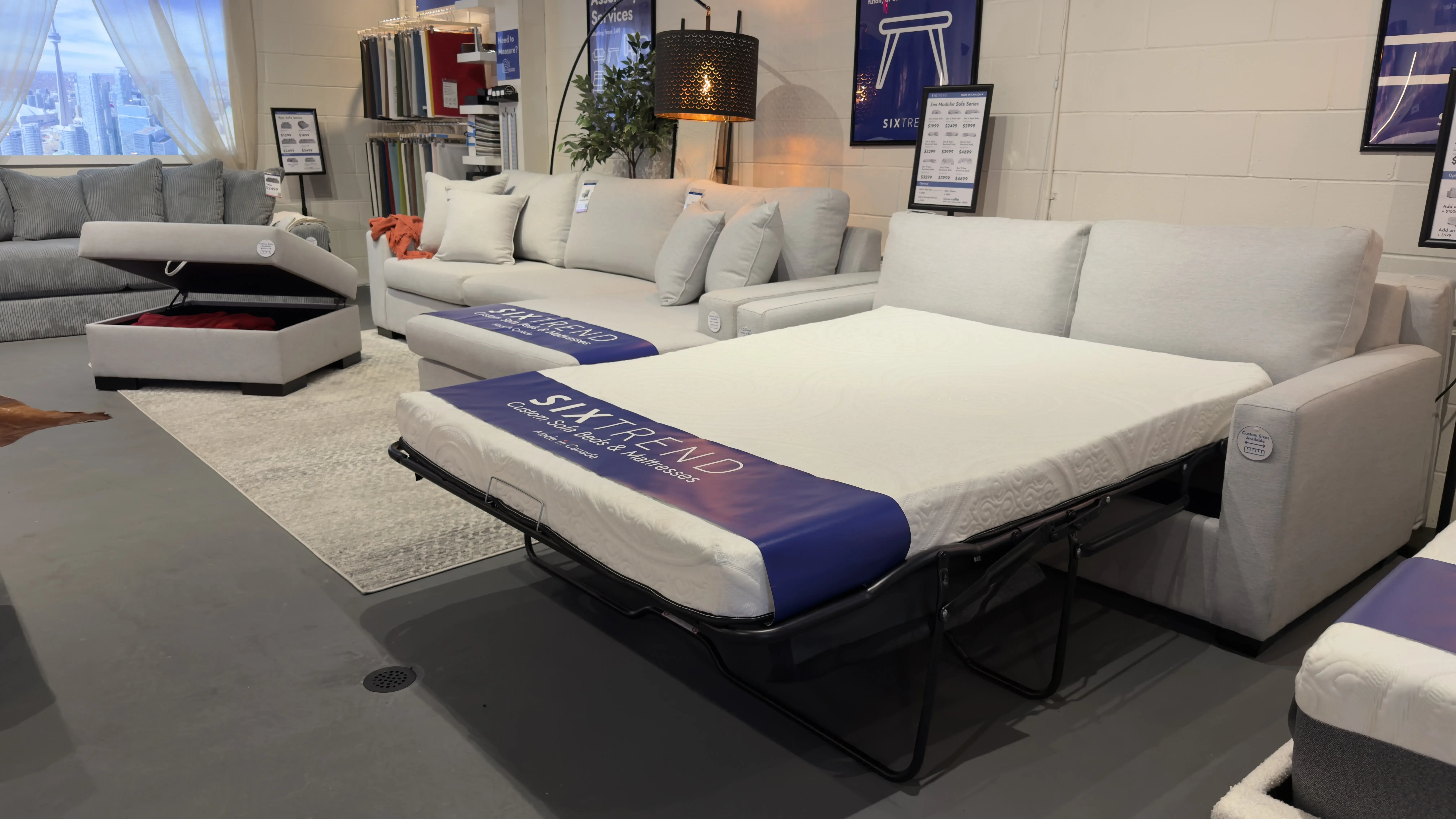 Light grey sofa bed with the queen mattress fully extended on its sleeper frame in a SIXTREND showroom, beside a matching sofa with storage ottoman.