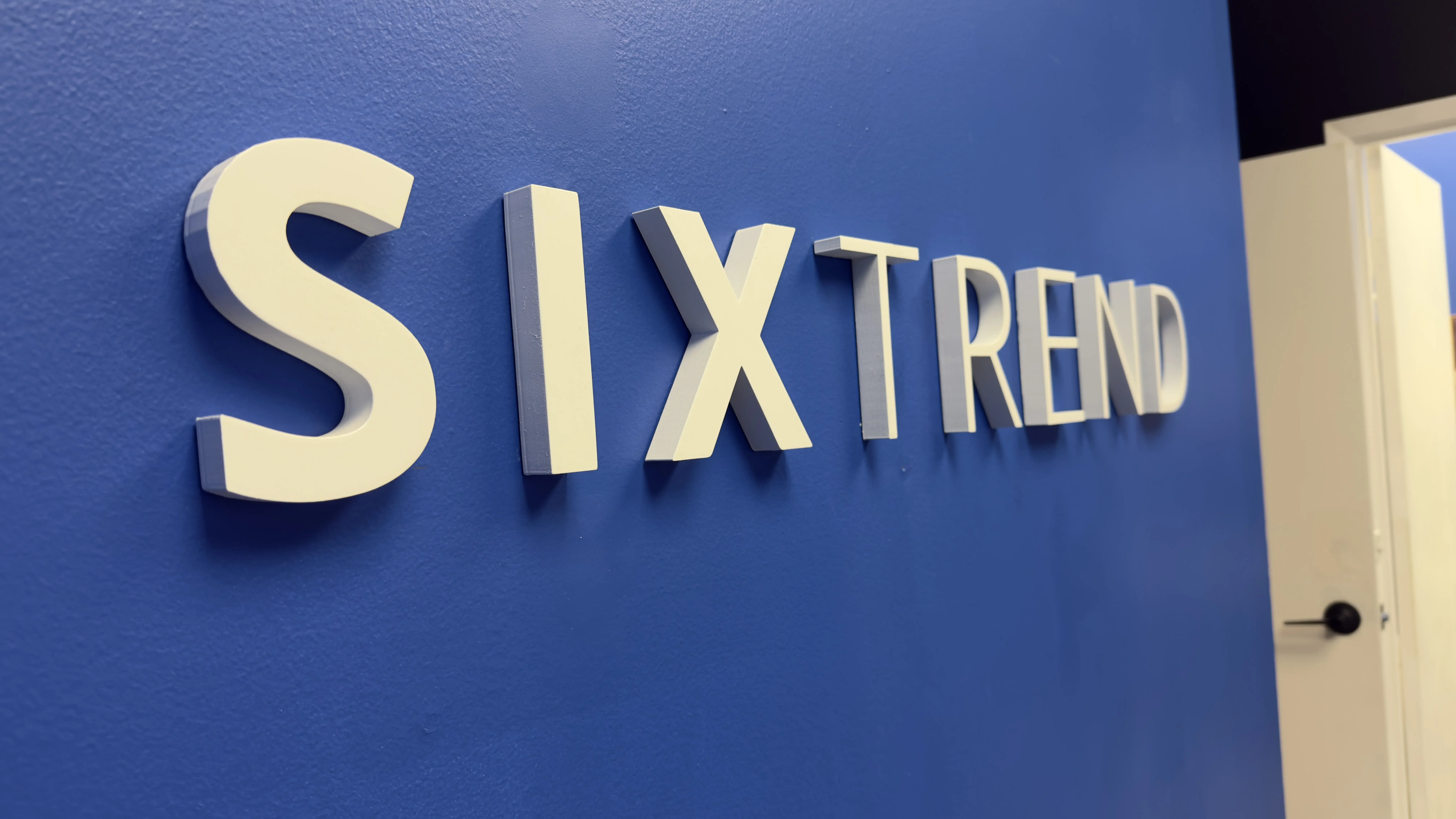 Three-dimensional white SIXTREND wordmark sign mounted on a deep blue accent wall in the company showroom.